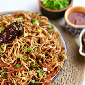 "A bowl of steaming noodles garnished with fresh vegetables, herbs, and a flavorful sauce, served as a delicious and comforting dish."