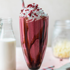 "A decadent Red Velvet Shake, featuring a vibrant red color, topped with a swirl of whipped cream and a drizzle of rich cream cheese frosting. The shake is served in a tall glass, garnished with red velvet cake crumbs and a cherry on top, making it a perfect indulgent treat."
